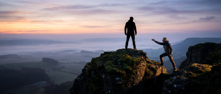 two men on mountain tops, one reaching out to the other. trainer confidence building at sunset.