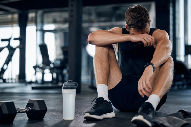 sweaty man in gym sitting on floor, head in hands, next to dumbbell and water bottle to prevent training stagnation.