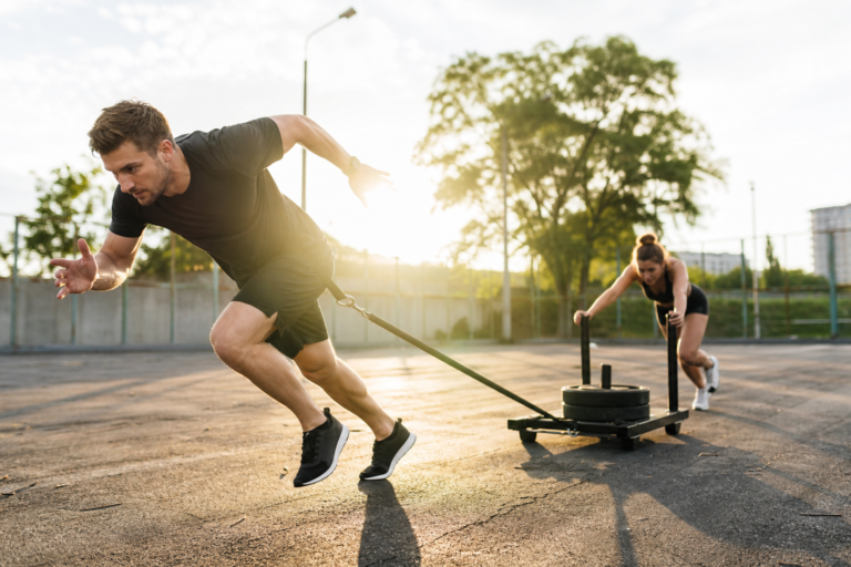 man pulling a weight sled and woman pushing another, showing personal training for performance gains, outdoors at sunset.