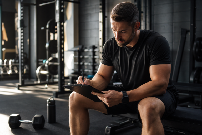 man on bench writing workout plan, ensuring returning to gym safely with a fitness coach.